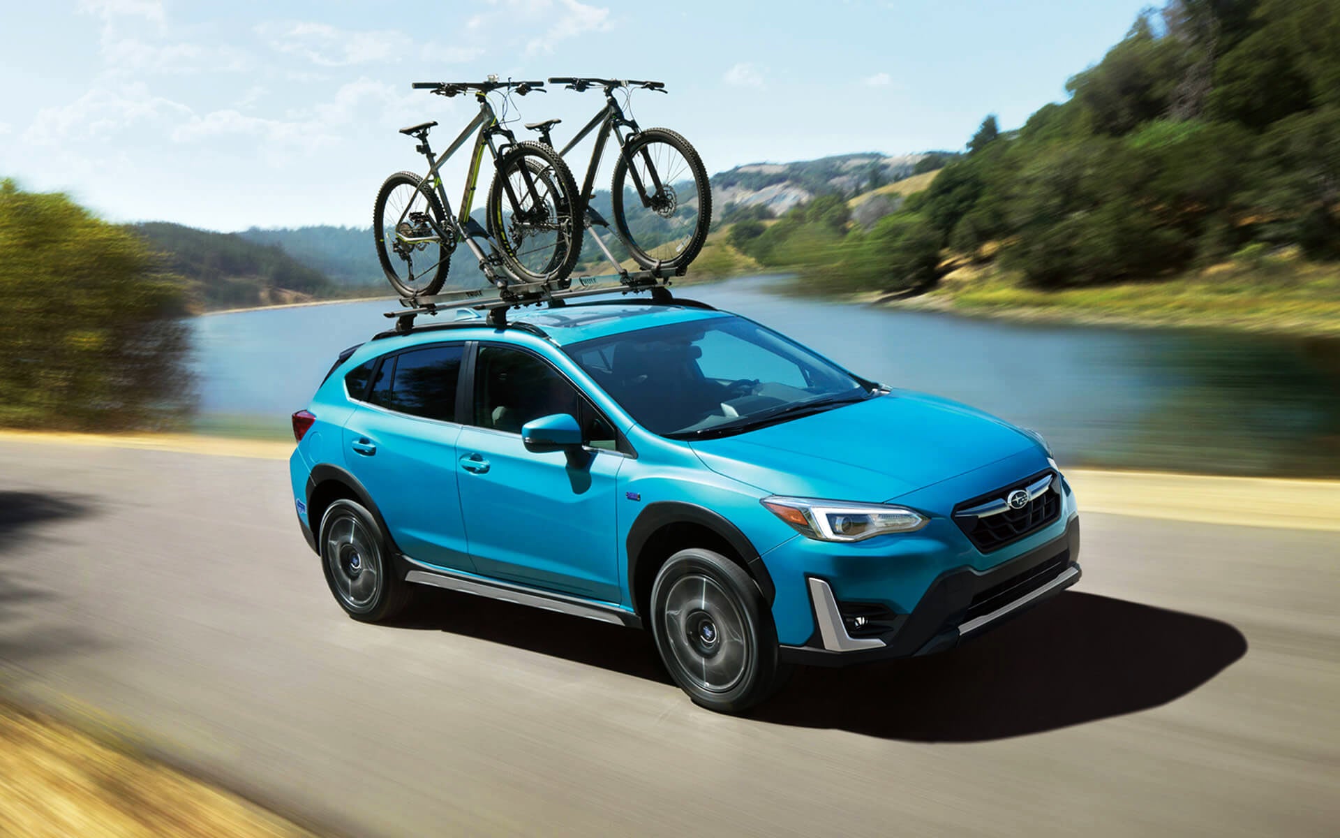 A blue Crosstrek Hybrid with two bicycles on its roof rack driving beside a river | Thelen Subaru in Bay City MI